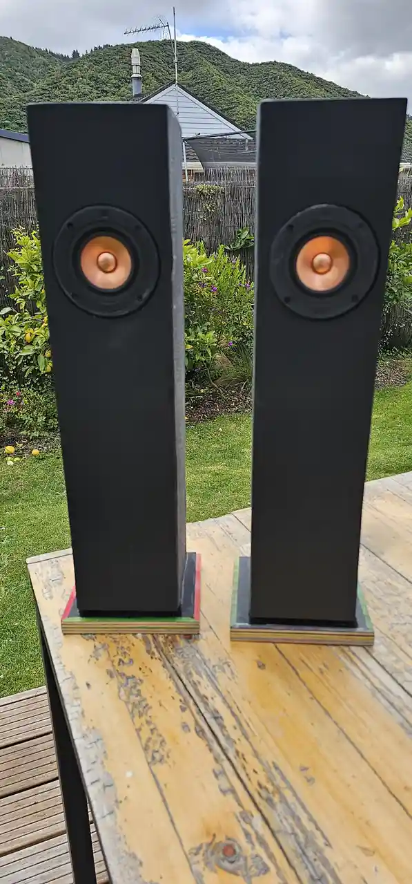 Two tall black floor-standing speakers with copper-colored cones sit on wooden stands outdoors on a deck, with a grassy…