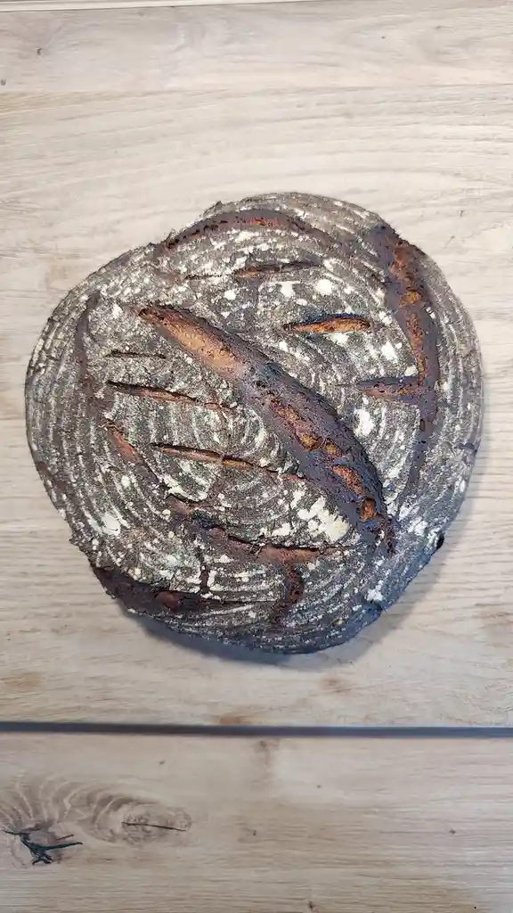 A round, rustic loaf of bread with a dark, cracked crust and flour dusting sits on a light wood surface. The bread's surface…