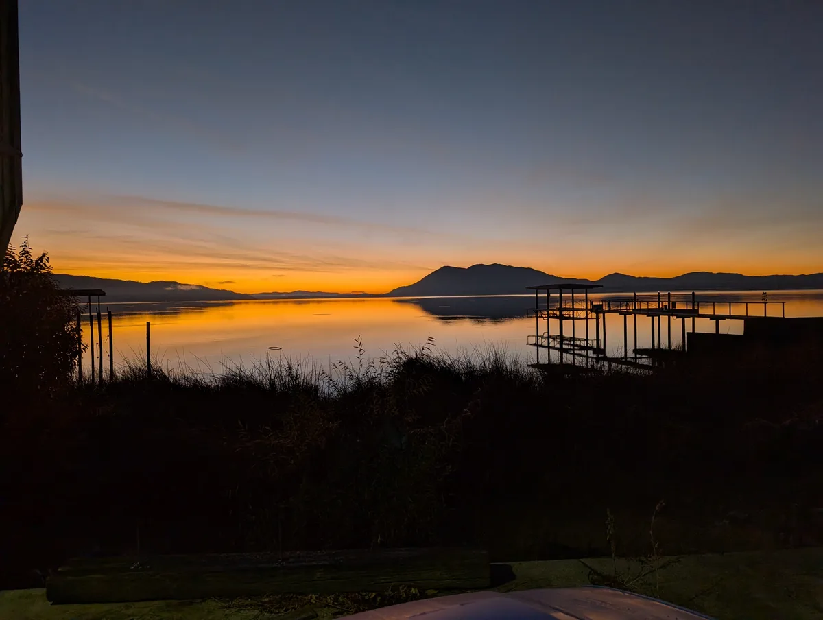 A serene sunset over a calm lake with distant mountains, featuring a wooden dock and silhouetted vegetation in the…