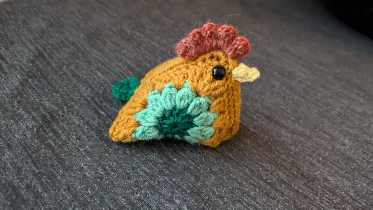 A small, hand-crocheted chicken figurine with a red comb, yellow beak, and black bead eye sits on a gray fabric surface. Its…