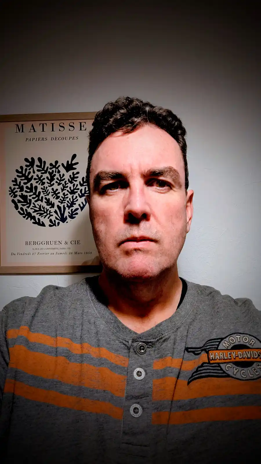 A man with a stern, unimpressed facial expression stares directly at the camera. He wears a grey and orange striped henley…
