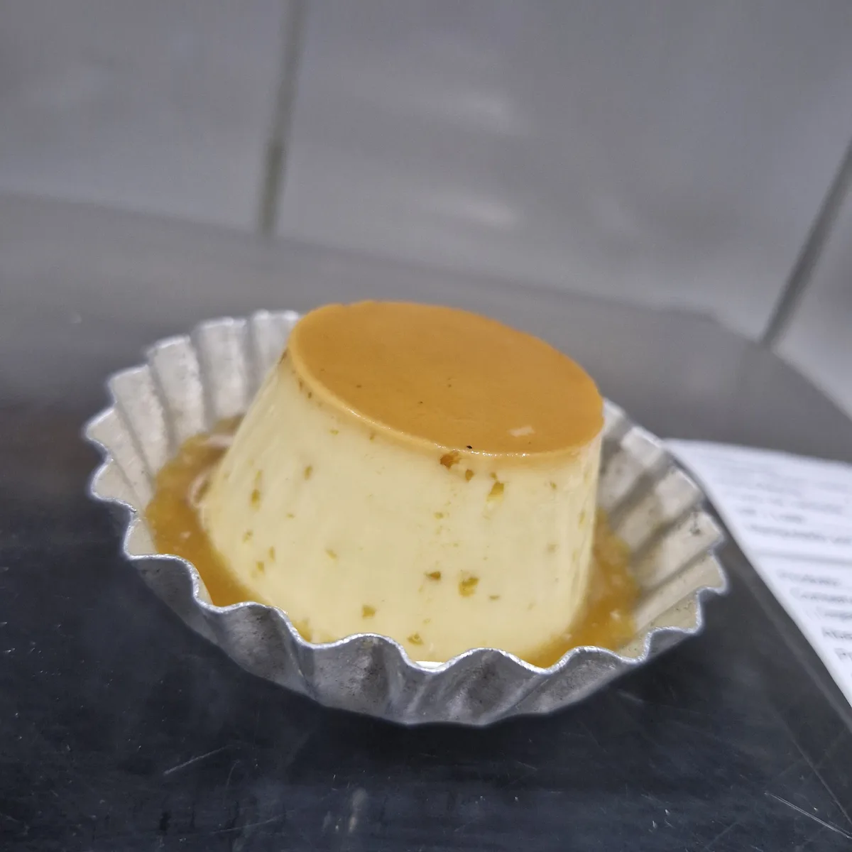 A close-up photo shows a small, perfectly cylindrical flan in a fluted metal baking mold, sitting on a dark countertop. The…