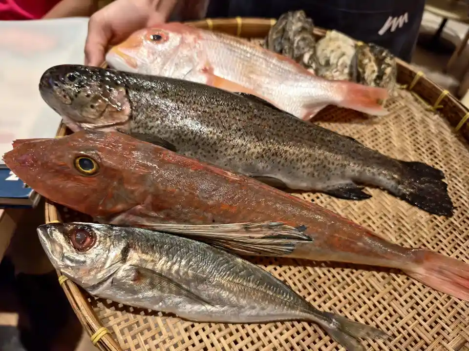 A close-up photo shows four different types of fresh fish and several oysters arranged on a woven bamboo tray. A person's…