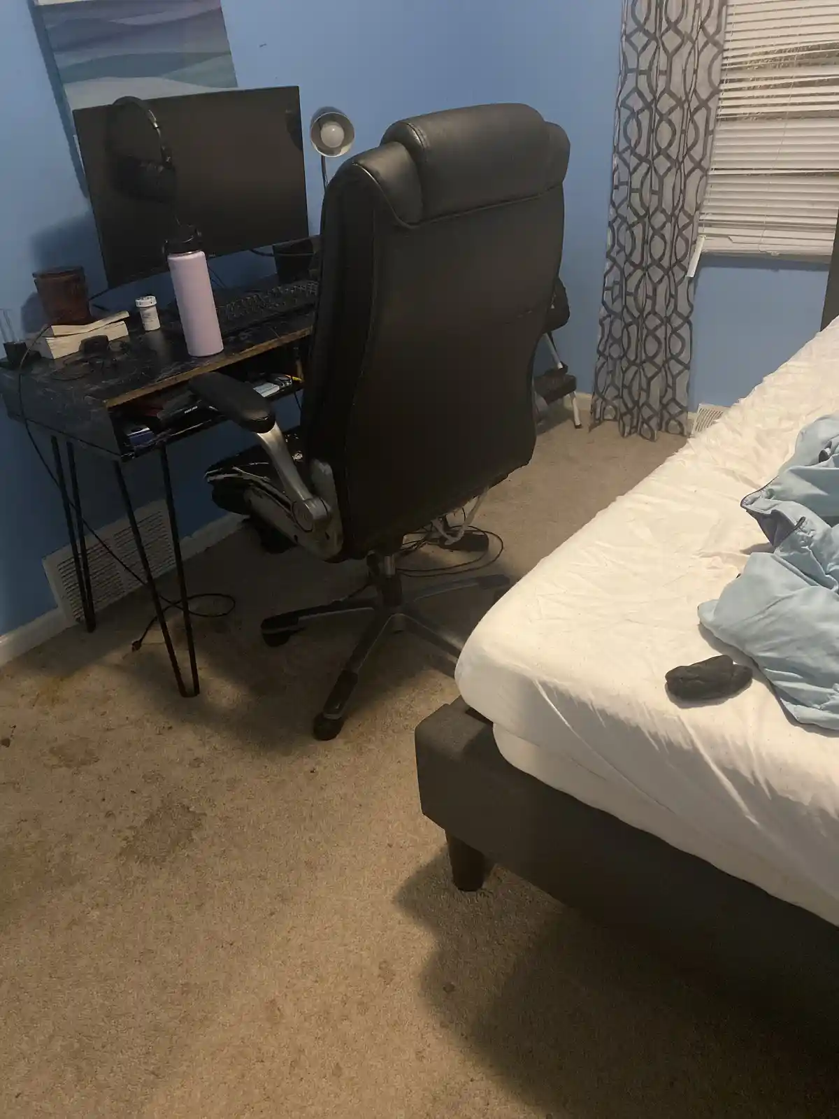 A bedroom with a black ergonomic office chair facing a desk with a monitor, headphones, and a water bottle. A bed with…