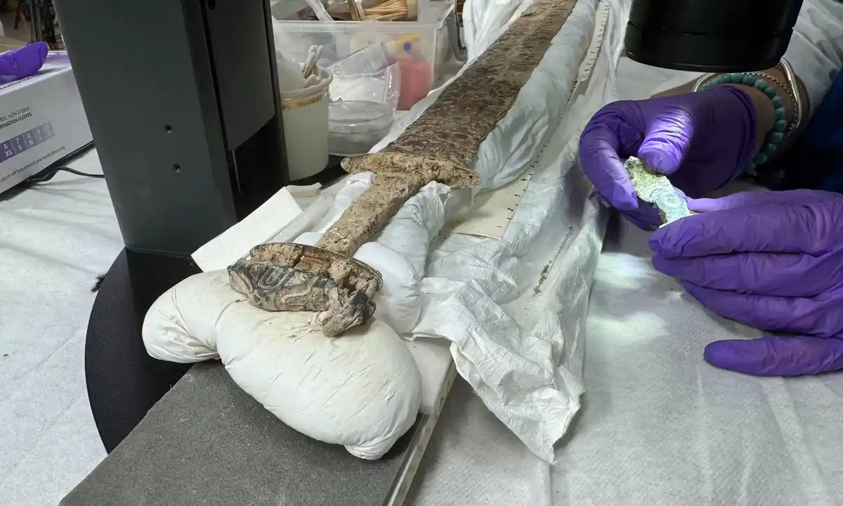 A person wearing purple gloves holds a crumpled dollar bill while a heavily corroded, ancient sword lies on a white cloth…