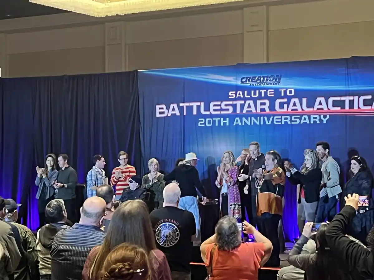 A group of people stand on a stage in front of a blue banner that reads 'Salute to Battlestar Galactica 20th Anniversary'…