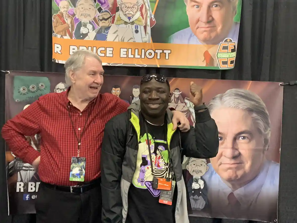 Two men pose for a photo at a comic con, standing in front of banners featuring Bruce Elliott and anime characters. The man…
