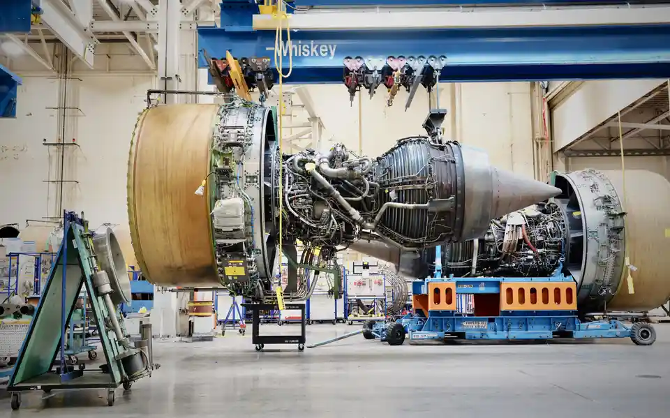 A massive, complex jet engine is suspended in an aircraft maintenance hangar by a blue overhead crane labeled 'Whiskey'. The…