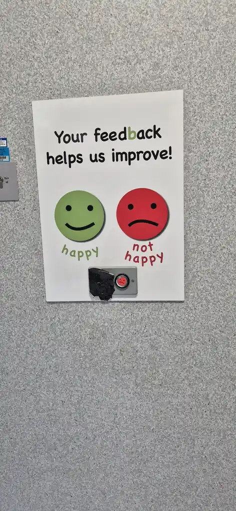 A white sign on a speckled grey wall displays the text 'Your feedback helps us improve!' above two circular icons: a green…