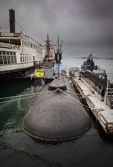 A black submarine is docked at a pier under a cloudy sky, with a yellow sign reading 'ENTRANCE' attached to its conning…