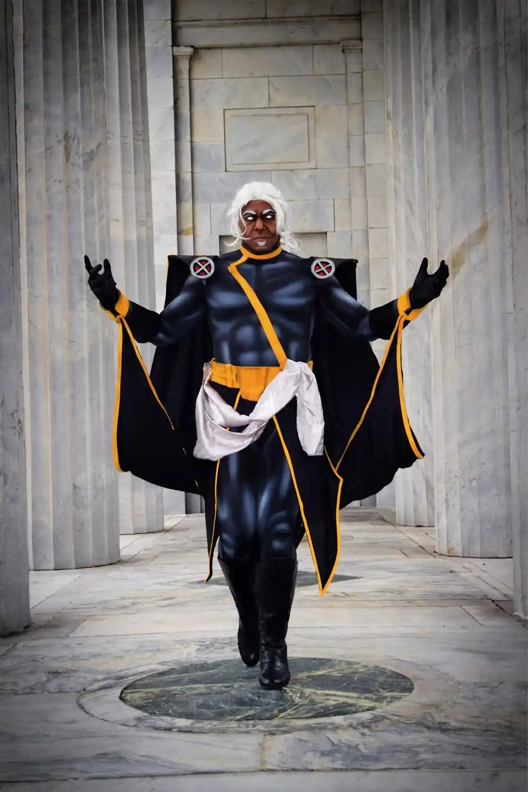 A person in a black and yellow X-Men costume with white wig and dramatic makeup stands in a grand marble hall with columns…