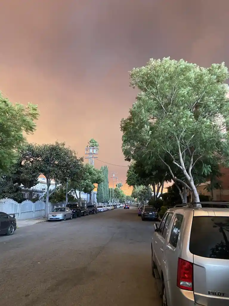 A residential street lined with parked cars and trees under a sky filled with thick, orange-brown smoke, making it appear as…