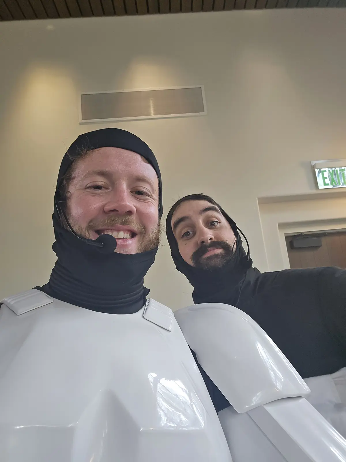 Two men in white stormtrooper armor with black balaclavas and headsets pose for a selfie indoors. The man on the left smiles…