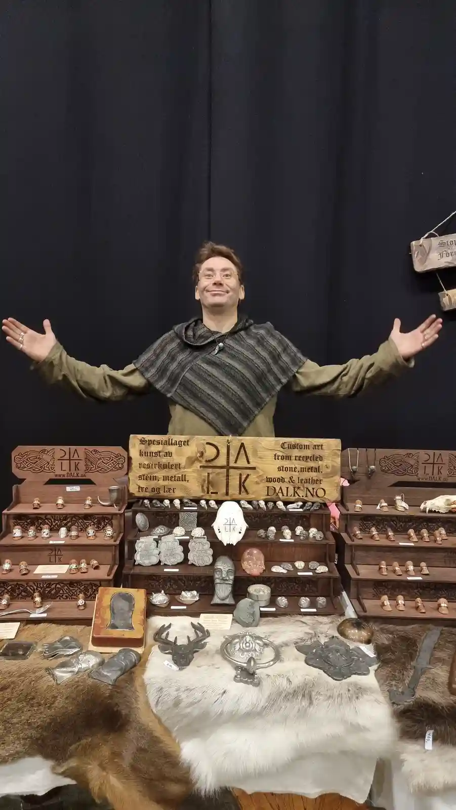 A man in medieval-style clothing stands behind a display table covered in animal furs, smiling with arms wide open. The…