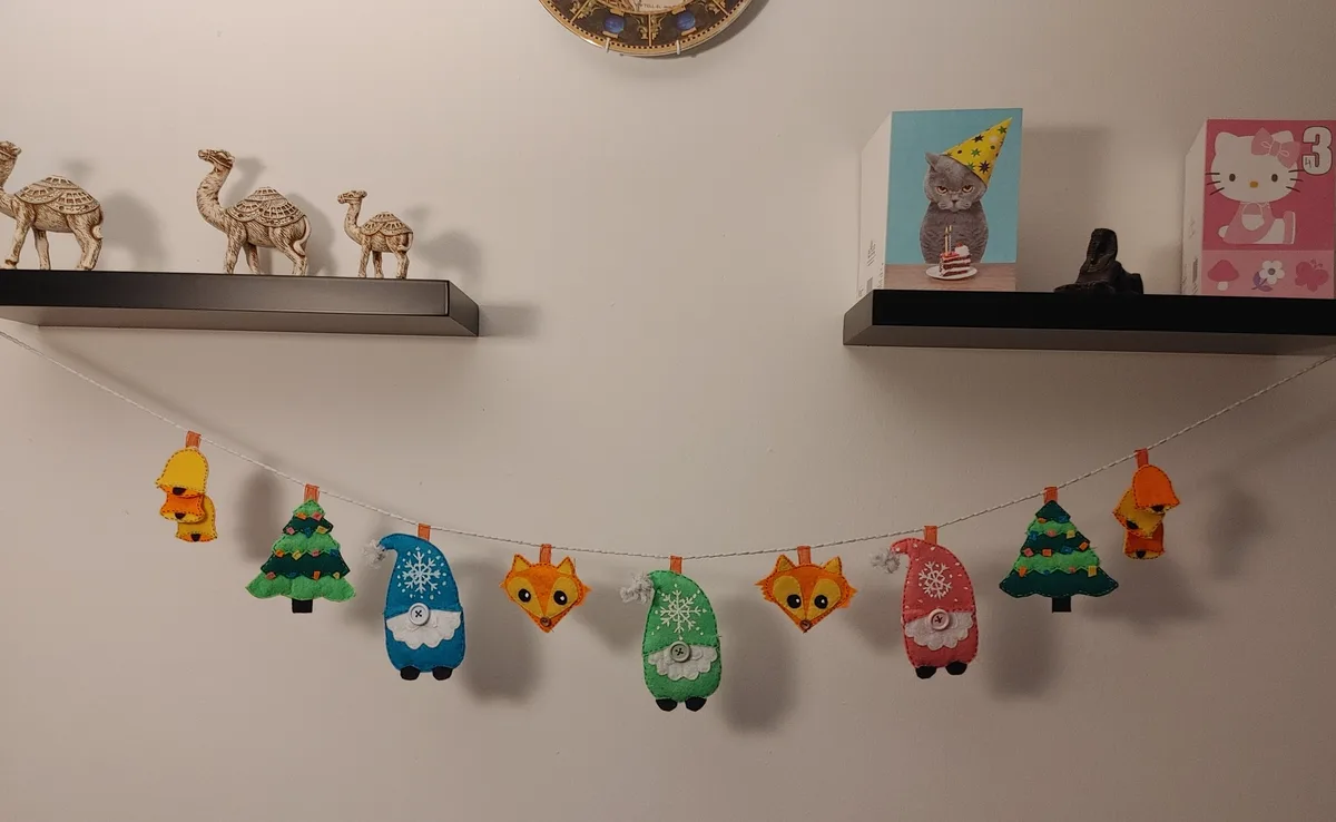 A handmade felt garland with festive decorations like Christmas trees, gnomes, and foxes hangs between two black shelves…
