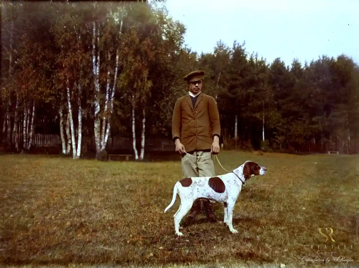 A vintage colorized photo shows a man in early 20th-century attire standing in a grassy field with a white and brown spotted…
