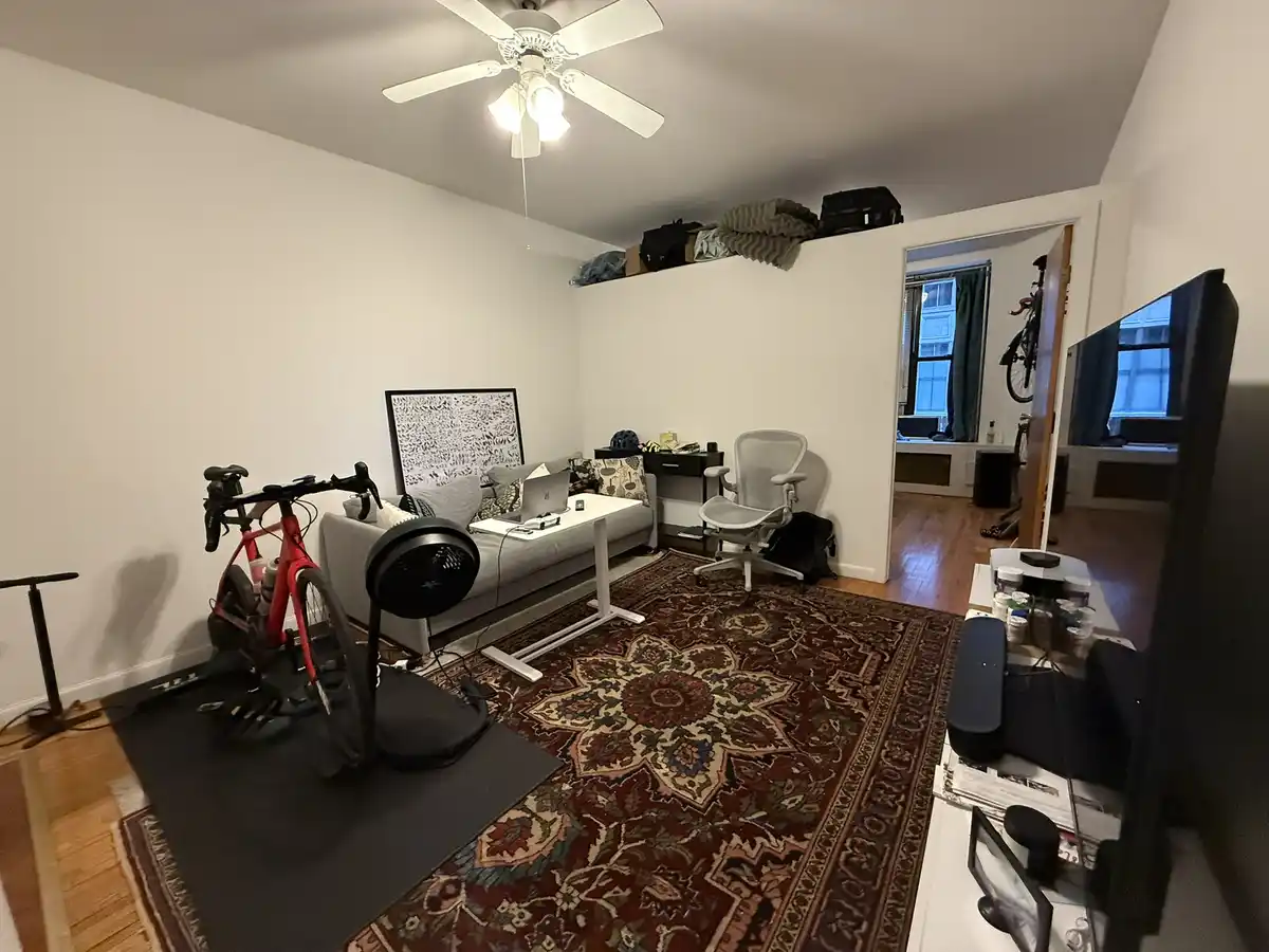 A cluttered apartment living room with a red stationary bike on a mat, a large ornate rug, a couch with a laptop, an…