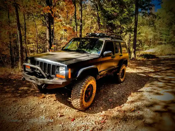 A black, lifted Jeep Cherokee with a roof rack and large off-road tires is parked on a gravel path in a forest with autumn…