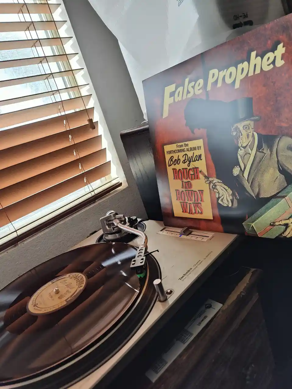 A vinyl record spins on a Technics turntable near a window with blinds, next to a poster for Bob Dylan's album 'Rough and…