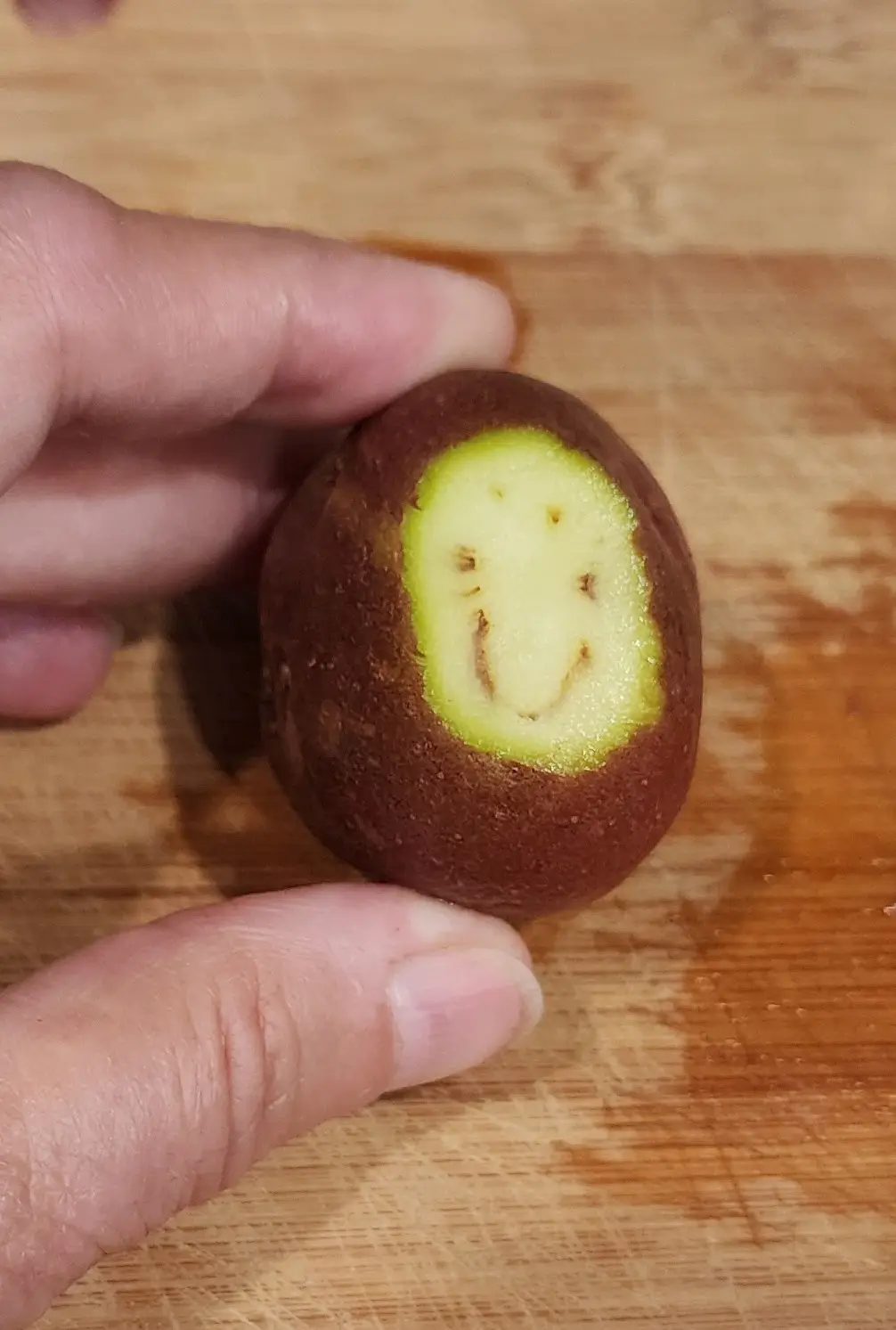 A hand holds a small, brown-skinned potato over a wooden cutting board, revealing a pale yellow interior with a natural…