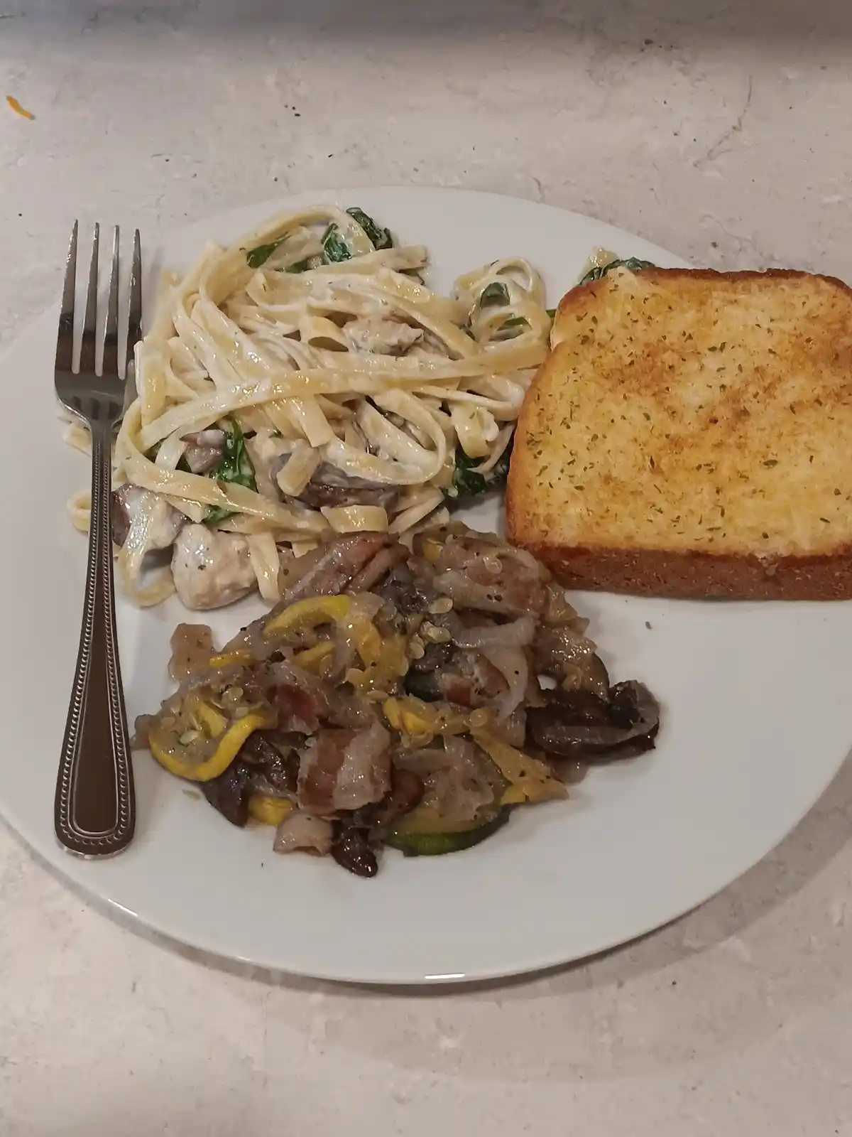 A white plate holds a meal with creamy fettuccine pasta with chicken and spinach, a slice of garlic bread, and a side of…
