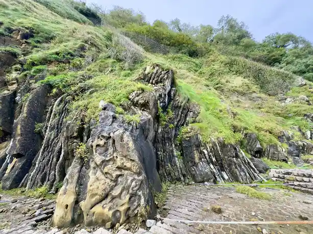 A photo of a steep, grassy cliffside with dark, vertically fractured rock formations and a stone path at the bottom…