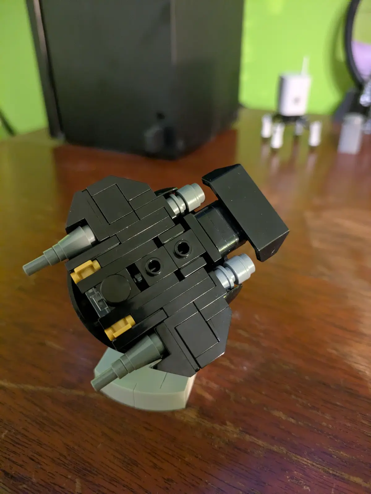 A close-up photo shows a black Lego spaceship model, viewed from the rear, sitting on a light gray baseplate atop a wooden…