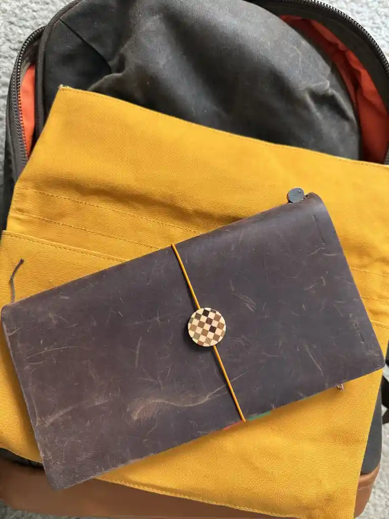 A close-up photo shows a well-worn, dark brown leather notebook with a checkered button closure resting on a mustard-yellow…