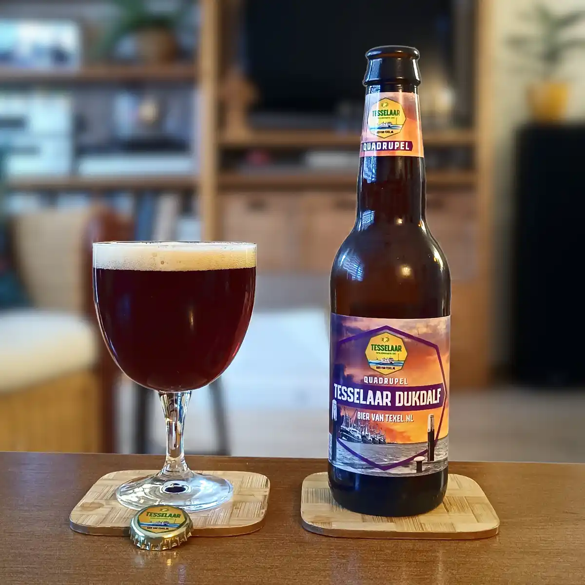 A bottle of Tesselaar Dukdalf Quadrupel beer and a filled glass sit on wooden coasters on a table, with the bottle cap…