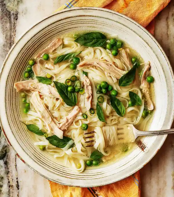 A top-down photo of a bowl of chicken noodle soup with peas and basil, with a fork resting in it. The overlaid text reads…