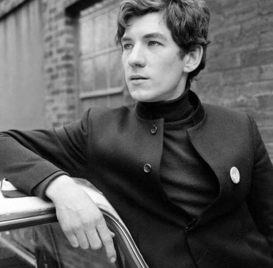 A black-and-white photo of a young man with dark hair, wearing a dark turtleneck and a dark coat with a circular pin on the…