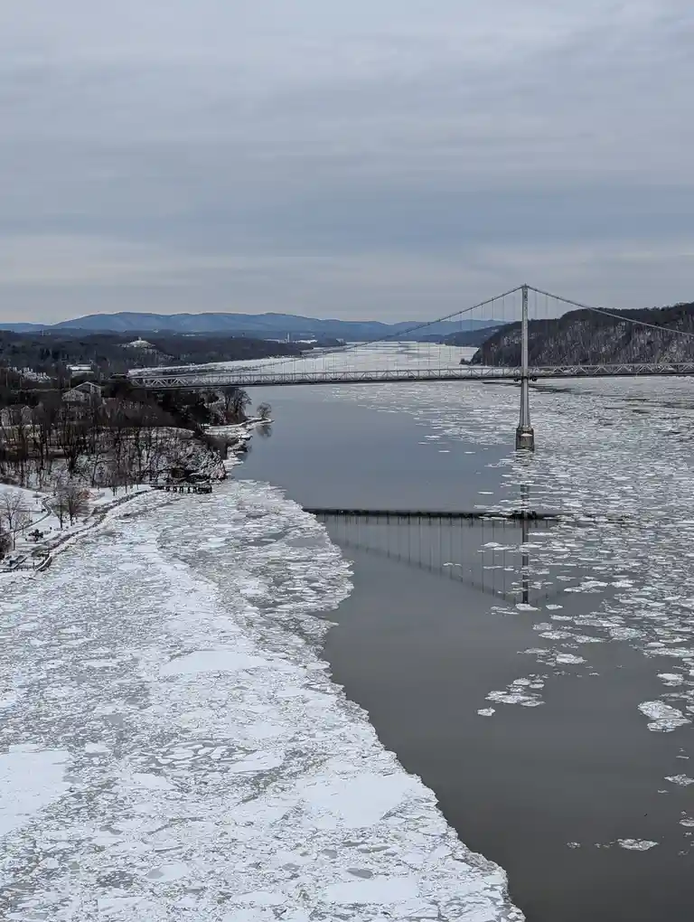A wide winter landscape shows a large suspension bridge spanning a partially frozen river, with ice floes floating in the…