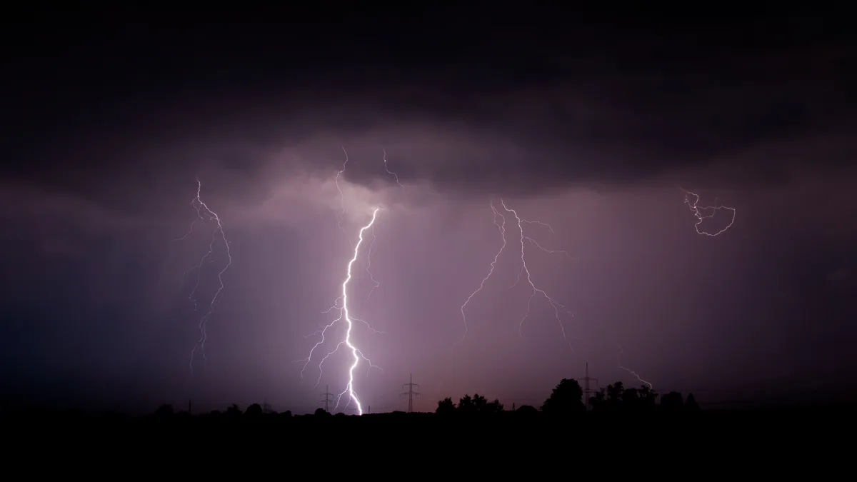 A dramatic night sky filled with multiple bright lightning bolts striking down from dark, stormy clouds over a silhouette of…