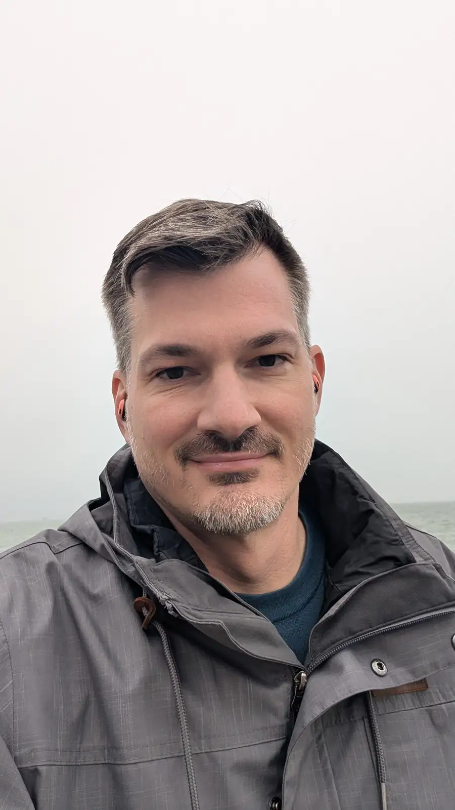 A man with a gray jacket and earbuds smiles at the camera against a foggy, overcast backdrop with water visible in the…