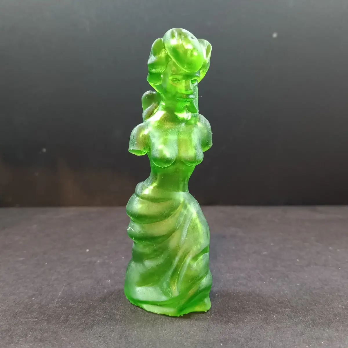 A translucent, bright green 3D-printed figurine of a woman with a stylized, retro hairstyle and draped gown stands against a…