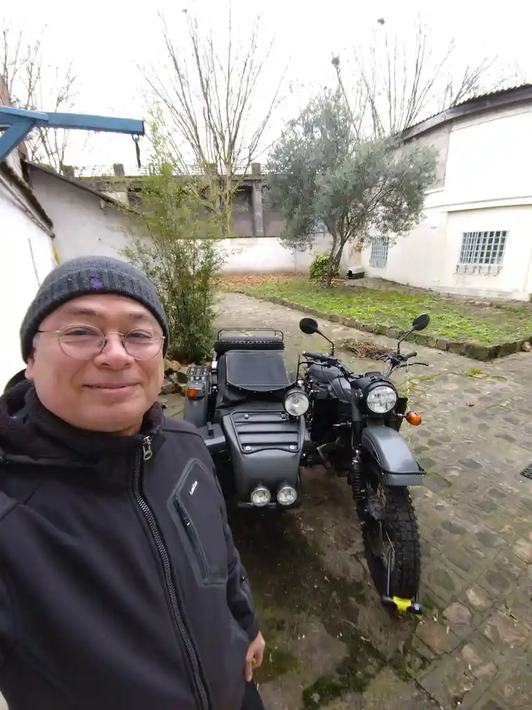 A man in a grey beanie and glasses smiles at the camera while standing beside a black motorcycle with a matching sidecar…