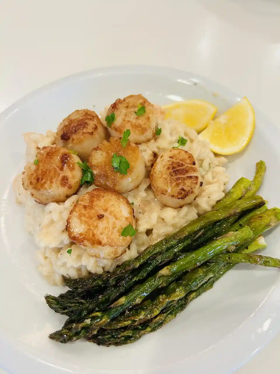 A close-up photo of a plated meal featuring seared scallops atop creamy risotto, accompanied by roasted asparagus spears and…