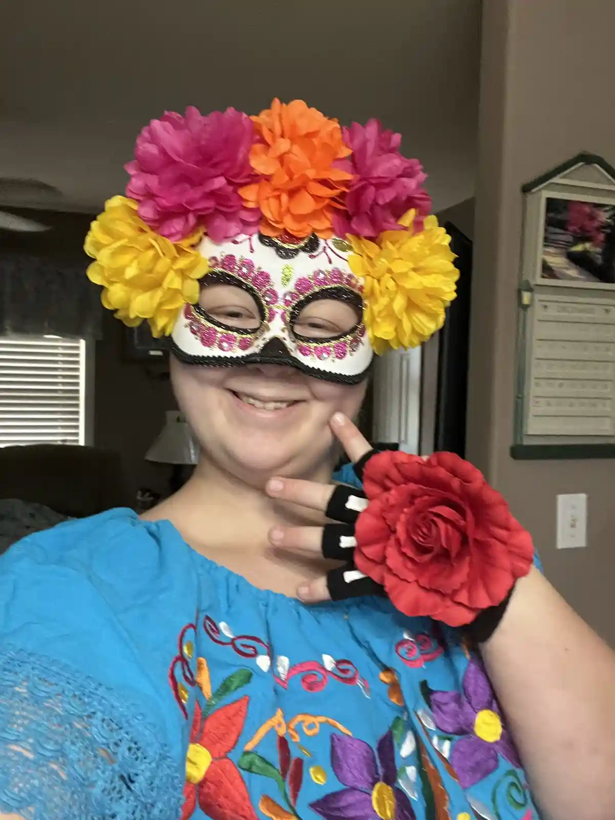 A person wearing a colorful sugar skull mask adorned with large pink, orange, and yellow paper flowers, paired with a blue…