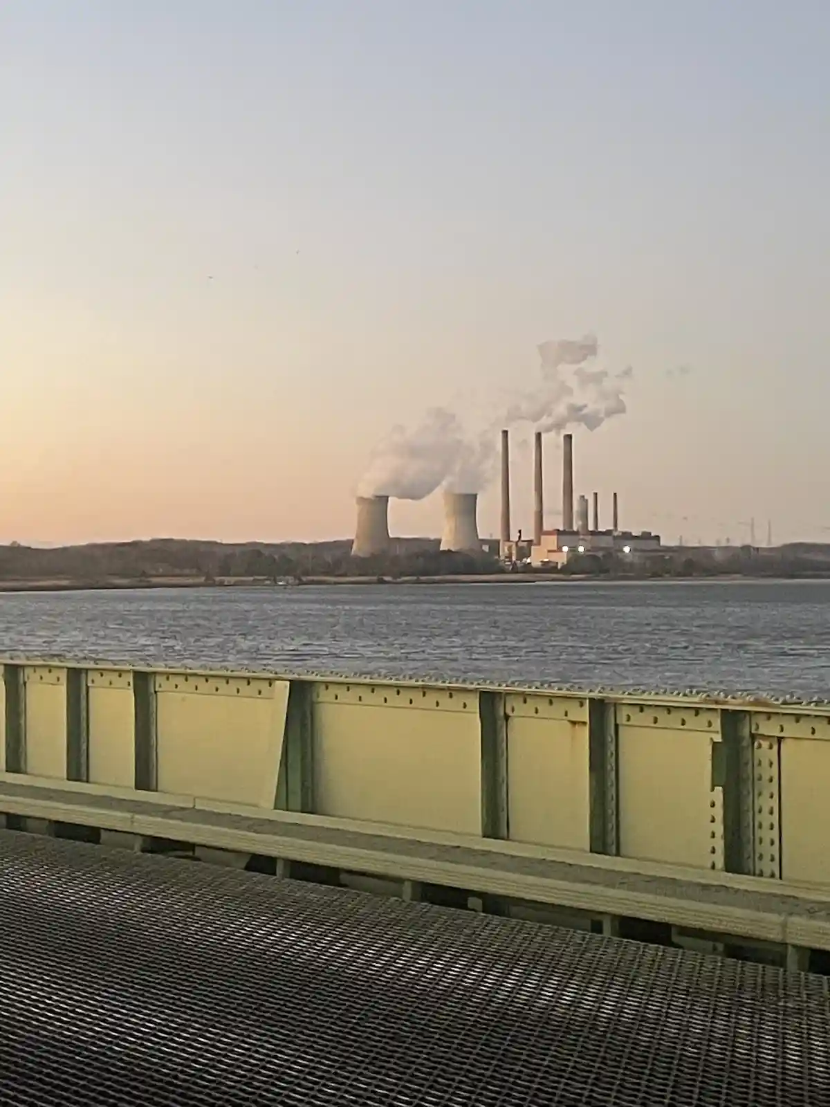 A photo taken from a bridge shows a large power plant with cooling towers and smokestacks emitting plumes of smoke over a…
