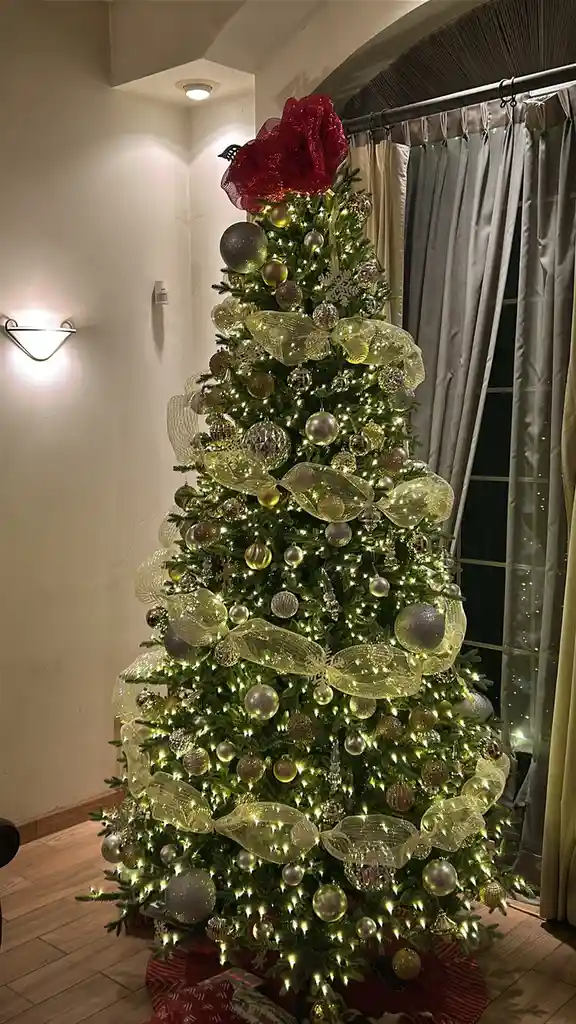 A lavishly decorated Christmas tree with gold and silver ornaments, glowing lights, and large sheer bows stands in a room…