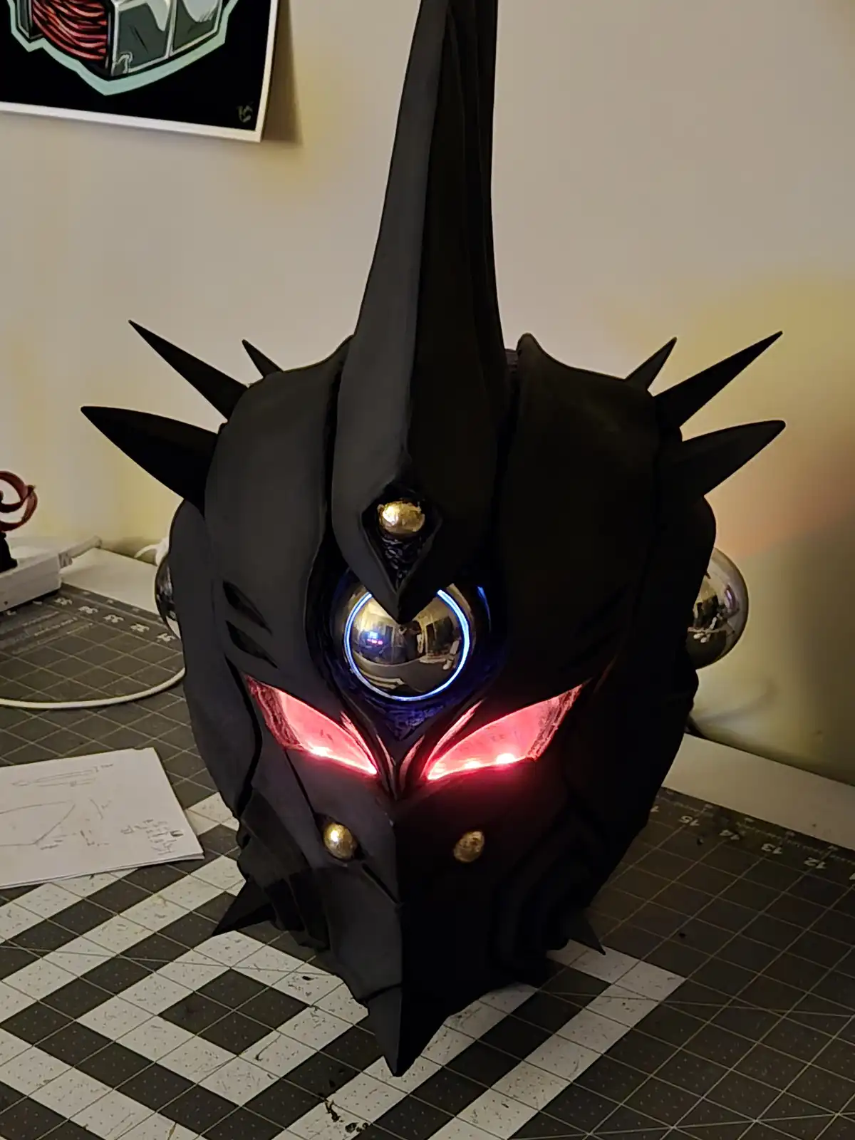 A black, spiky, angular cosplay mask with glowing red eyes and a central blue-lit orb sits on a checkered crafting mat. The…