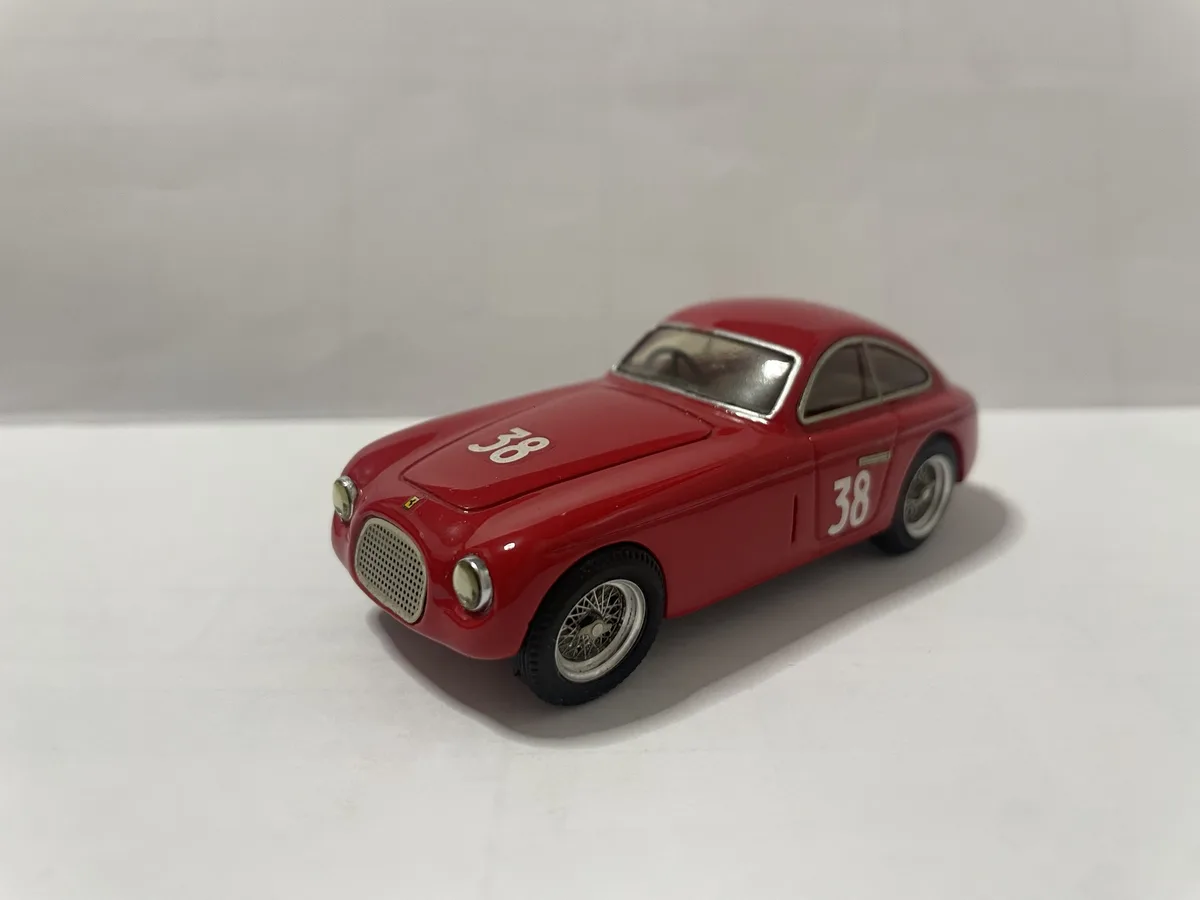 A red die-cast model of a vintage Ferrari race car, marked with the number 38 on its hood and side, sits on a plain white…