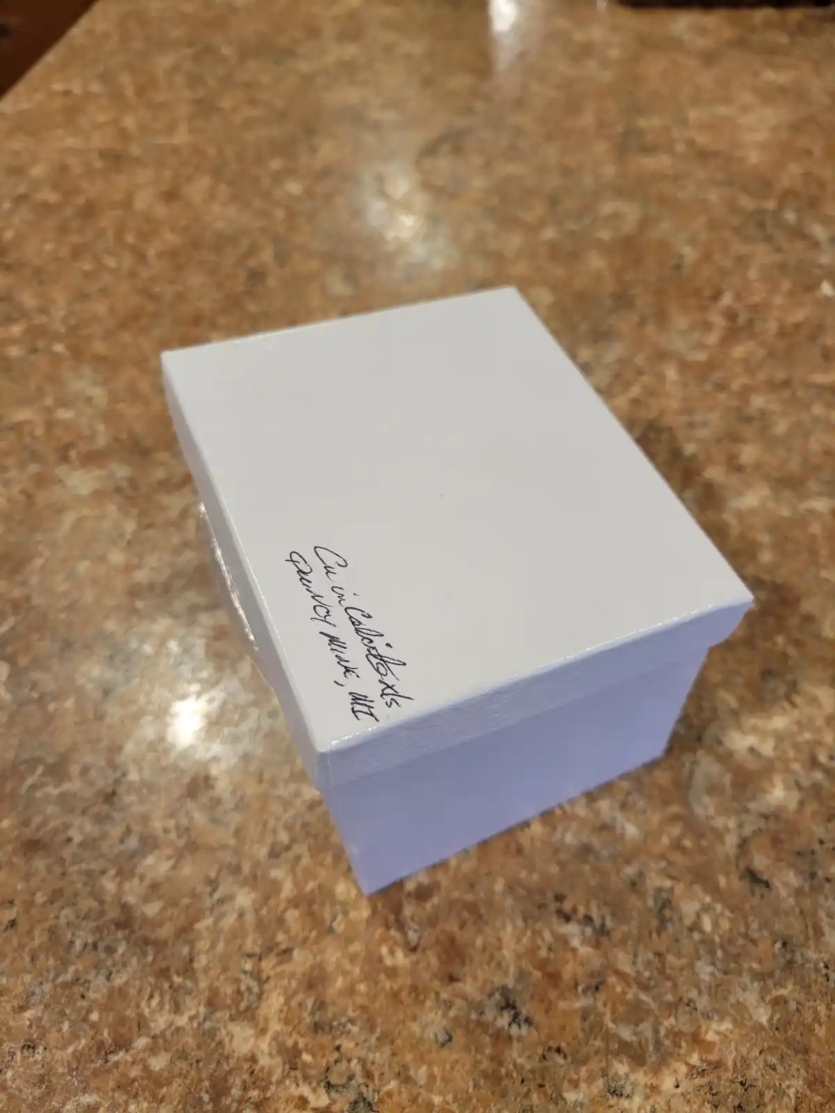 A plain white rectangular box sits on a speckled brown countertop, with handwritten text on its lid that reads 'Cu in…