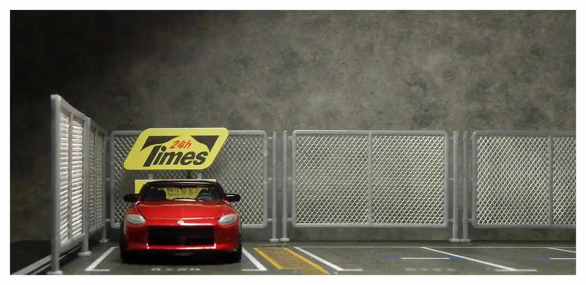 A red toy sports car is parked in a miniature parking lot with white lines and a yellow accessibility strip. Behind the car…