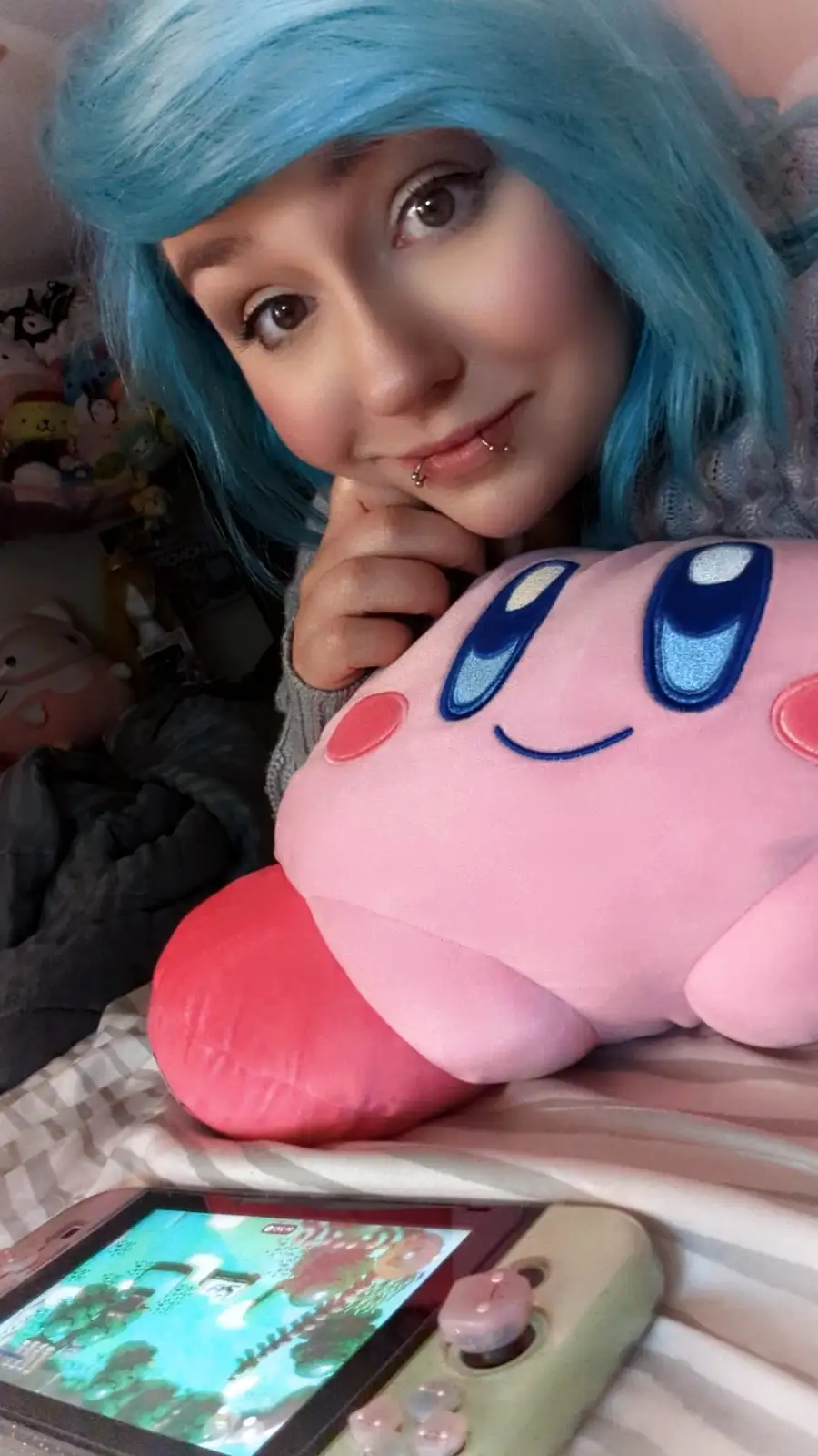 A person with blue hair and a lip piercing rests their chin on their hand, looking at the camera with a slight smile while…