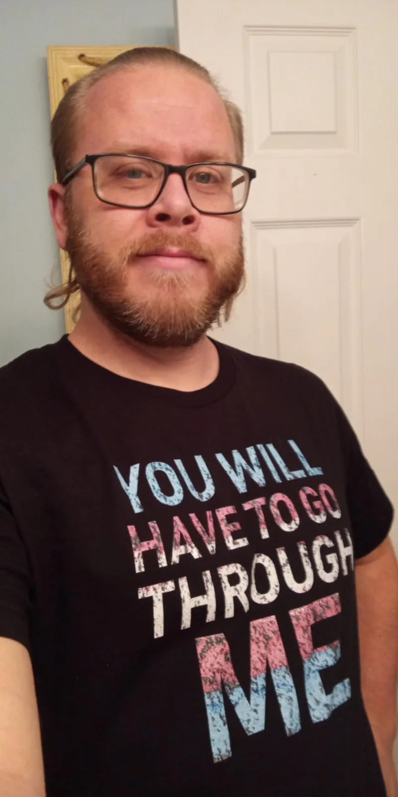 A man with glasses and a beard wears a black t-shirt with the text 'YOU WILL HAVE TO GO THROUGH ME' in a distressed…