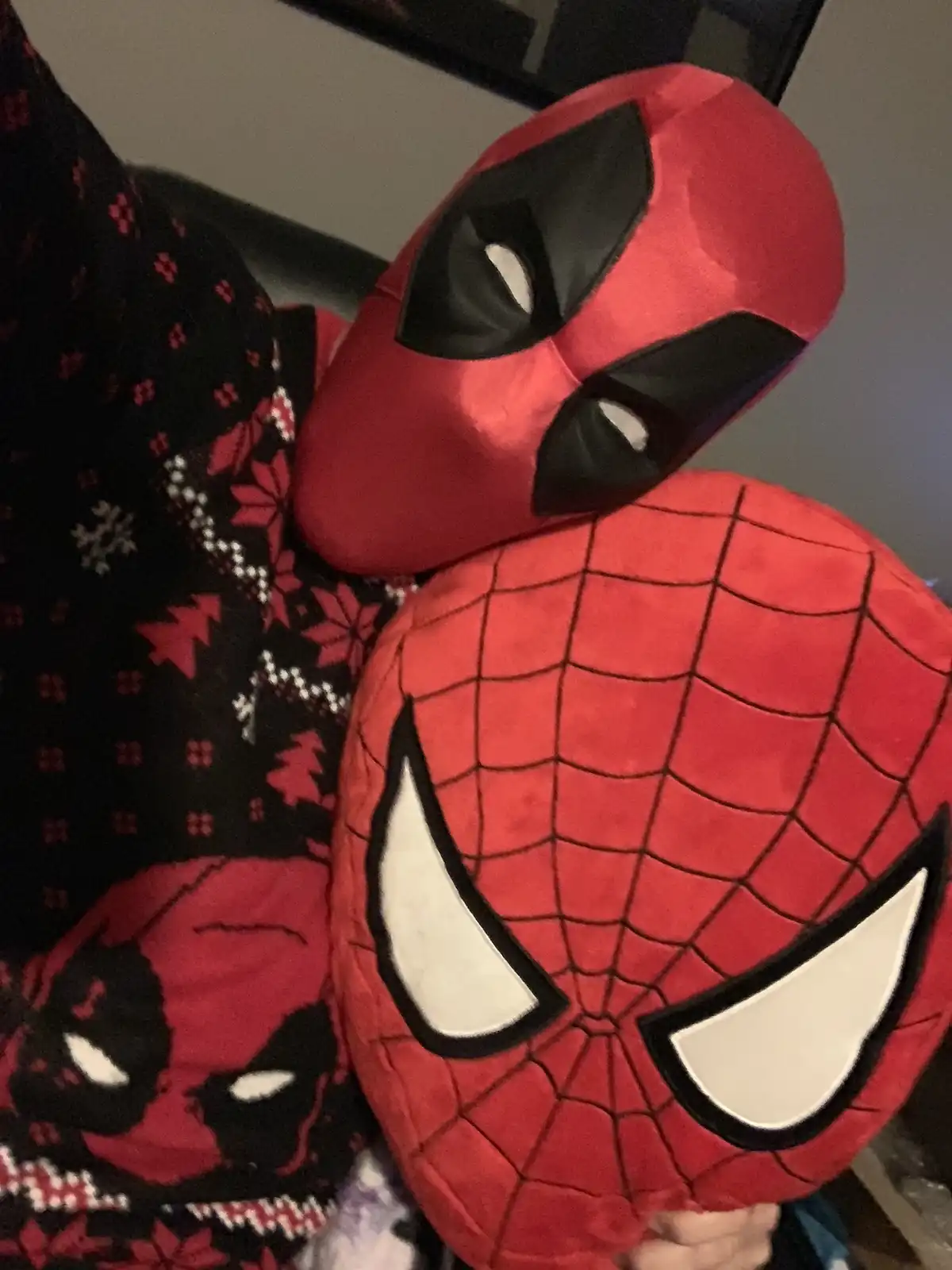 A red Deadpool plushie is hugging a red Spider-Man plushie, both resting against a black sweater with red and white…