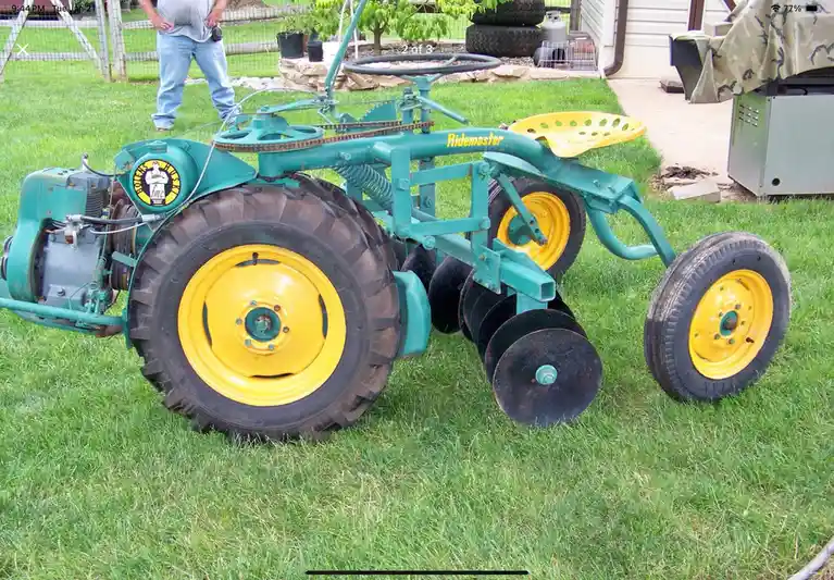 A teal and yellow Ridemaster garden tiller with large rear wheels and disc blades sits on a green lawn, with a person…