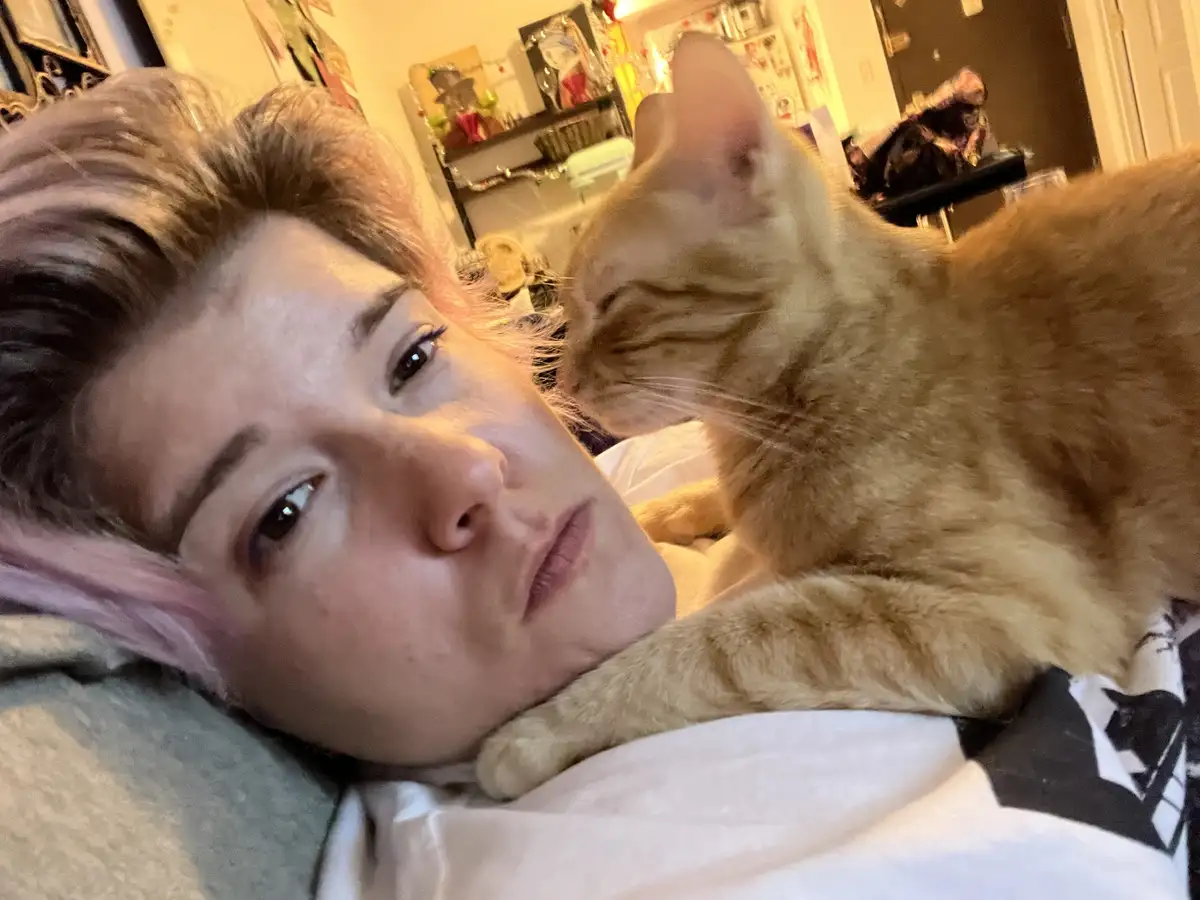 A person with pink-tipped hair lies in bed looking at the camera while an orange tabby cat rests its head and front paw on…