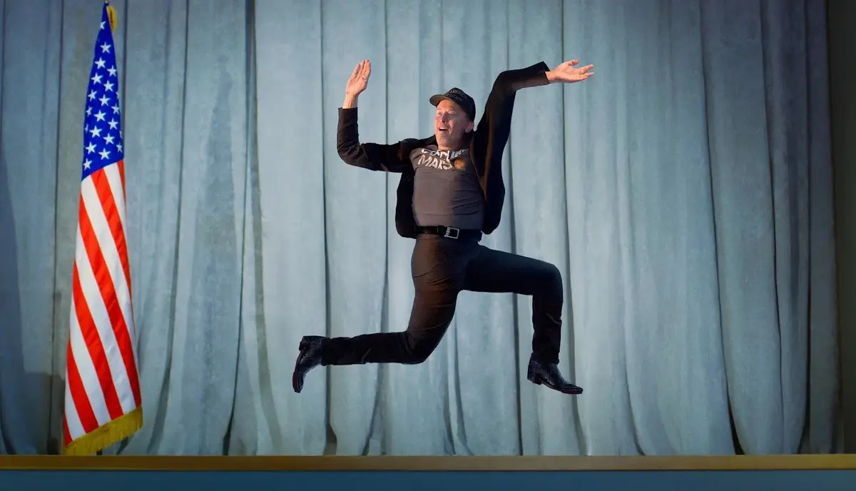 A man in a black jacket, grey t-shirt with 'COOL MARS' text, and a black cap is captured mid-air in a dynamic jumping pose…